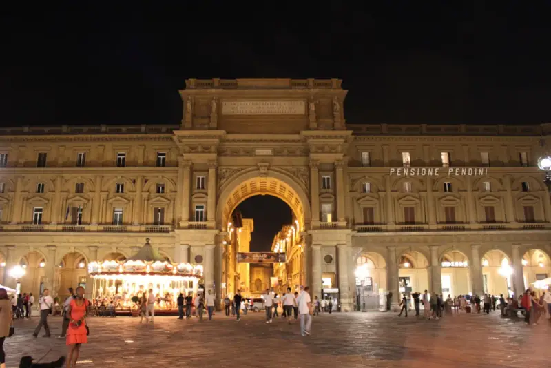 La piazza della Repubblica a Firenze di notte, con il suo arco monumentale illuminato, gente che passeggia e una giostra scintillante.