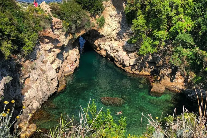 Bagni Regina Giovanna: una baia nascosta con acque cristalline, rocce scolpite e vegetazione lussureggiante. Persone si godono il sole e nuotano.