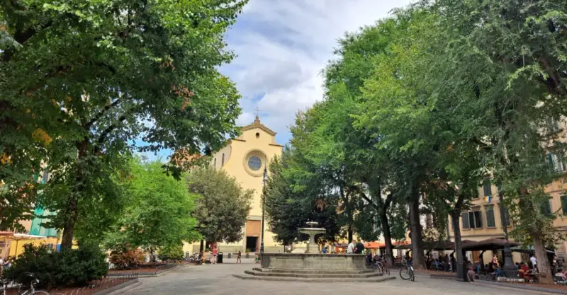 Piazza Santo Spirito a Firenze, con la chiesa sullo sfondo, una fontana al centro e alberi rigogliosi che incorniciano la scena.